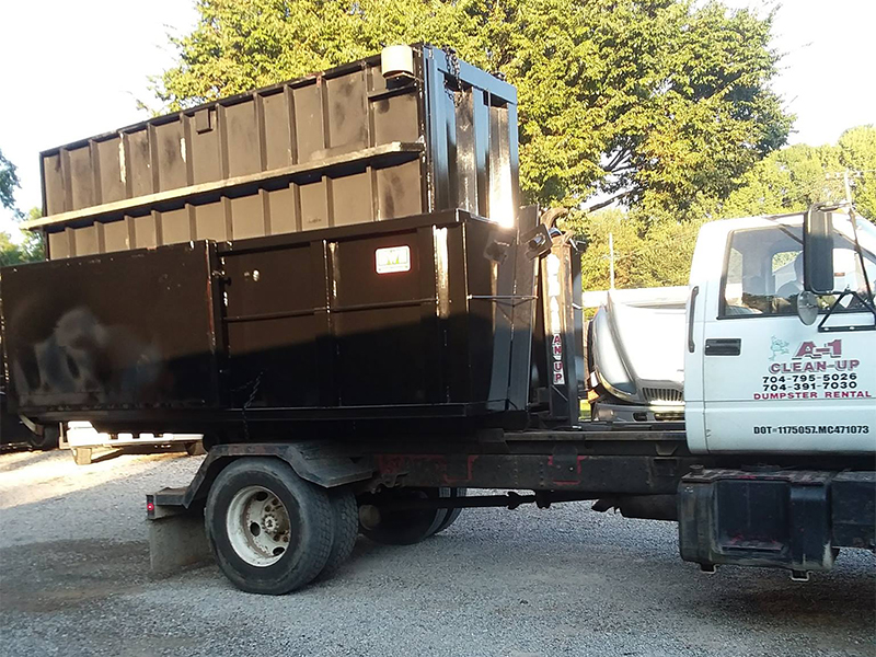 A black dumpster is loaded onto a flatbed truck in a gravel lot with trees in the background.